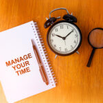 Notebook with the words MANAGE YOUR TIME in red with an alarm clock and magnifying glass sitting next to it.